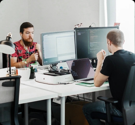 Dos ingenieros de QA trabajando en computadoras en una oficina de una empresa tecnológica, uno de ellos programando en dos monitores.