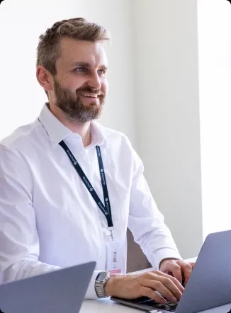 A man with a beard and white shirt sitting at the desk and smiling at a laptop.