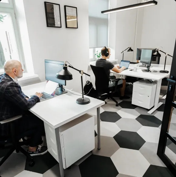 Zwei Männer arbeiten an getrennten Schreibtischen in einem modernen Büro, beide konzentriert auf ihre Laptops.