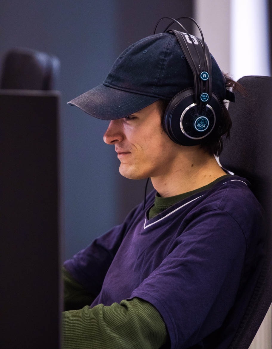 Laadunvarmistusinsinööri, jolla on lippis ja kuulokkeet, istuu työpöydän ääressä ja työskentelee pelitestiprojektissa.