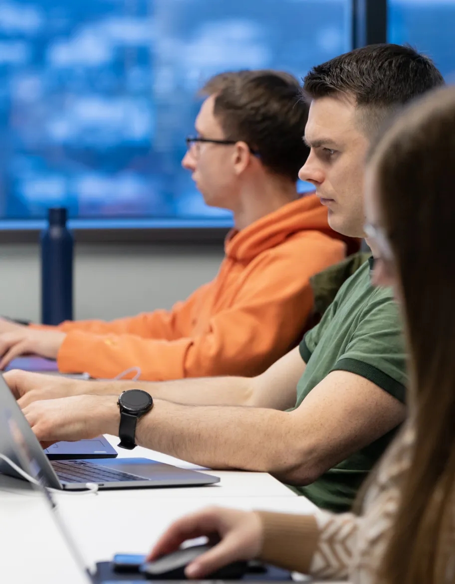 Kvalitātes nodrošināšanas inženieru grupa sēž pie rakstāmgalda ar portatīvajiem datoriem uz galda un piedalās sanāksmē.