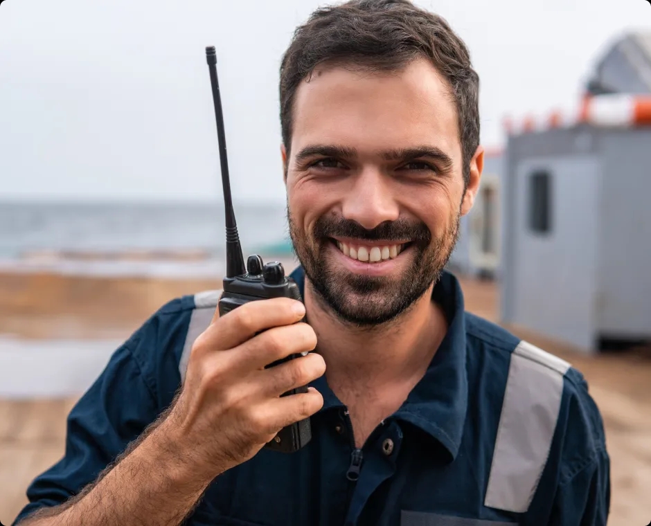 A man standing outdoors, holding a portable handheld radio device and smiling.