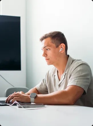 Eine Person sitzt an einem Schreibtisch, tippt auf einem Laptop, während ein großer Monitor im Hintergrund sichtbar ist.