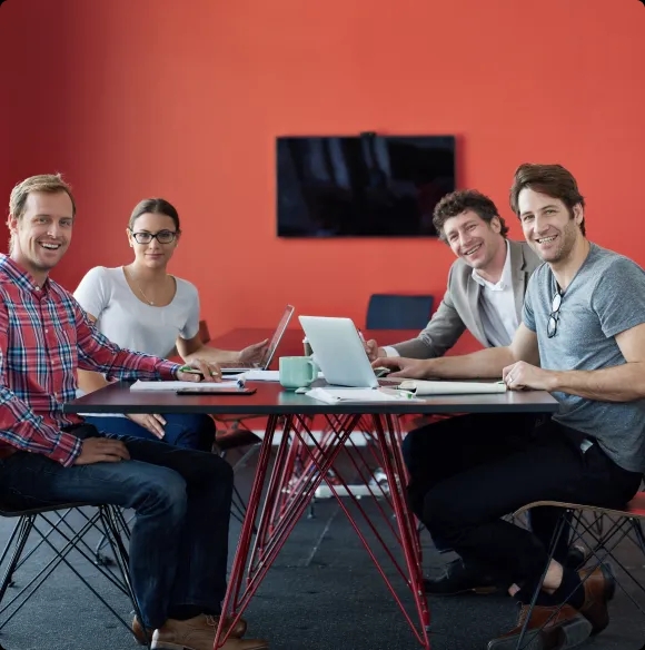 Four team members sitting at a table in a meeting room, all smiling and looking directly at the camera while working on laptops.