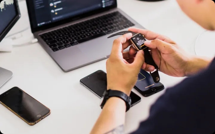 Hands holding a smartwatch next to a laptop on a desk.