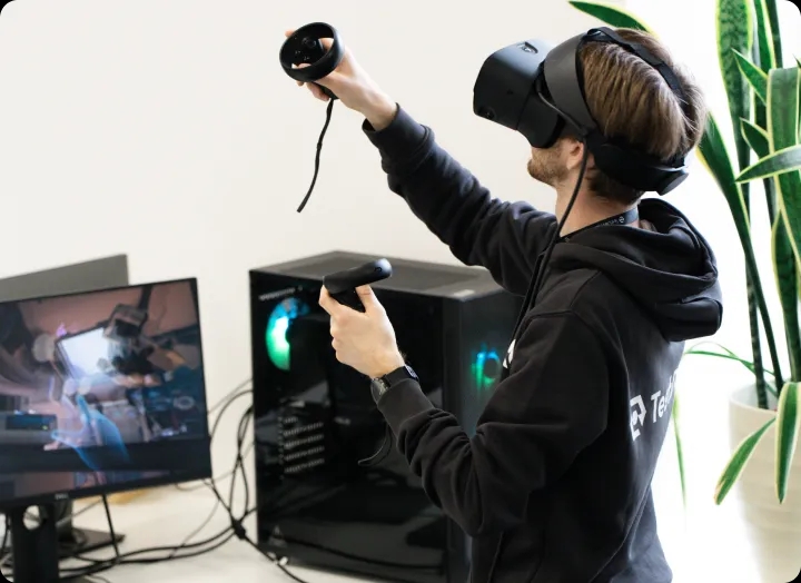 Eine Person steht und verwendet ein VR-Headset mit Controllern in der Nähe eines Computers.