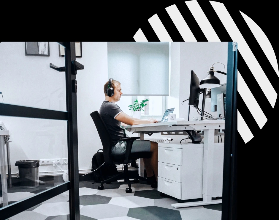 QA engineer working at a desk in an office, wearing headphones while focused on a laptop and monitor setup.