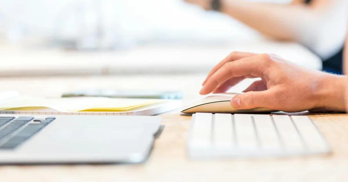 A hand interacting with a mouse on a desk with a laptop and keyboard in the foreground