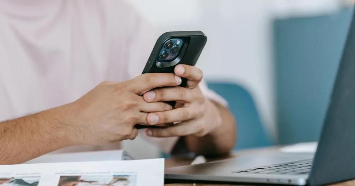 Person holding and using mobile phone while sitting at desk with a laptop