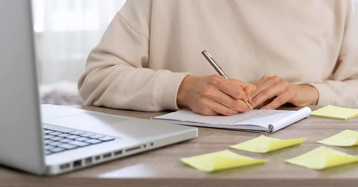 Person sitting at desk with laptop and writing in notebook