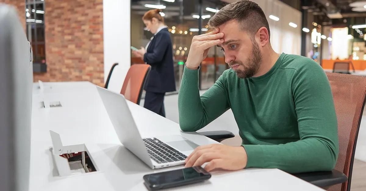 A QA engineer sitting at desk in office working on a laptop and looking fatigued