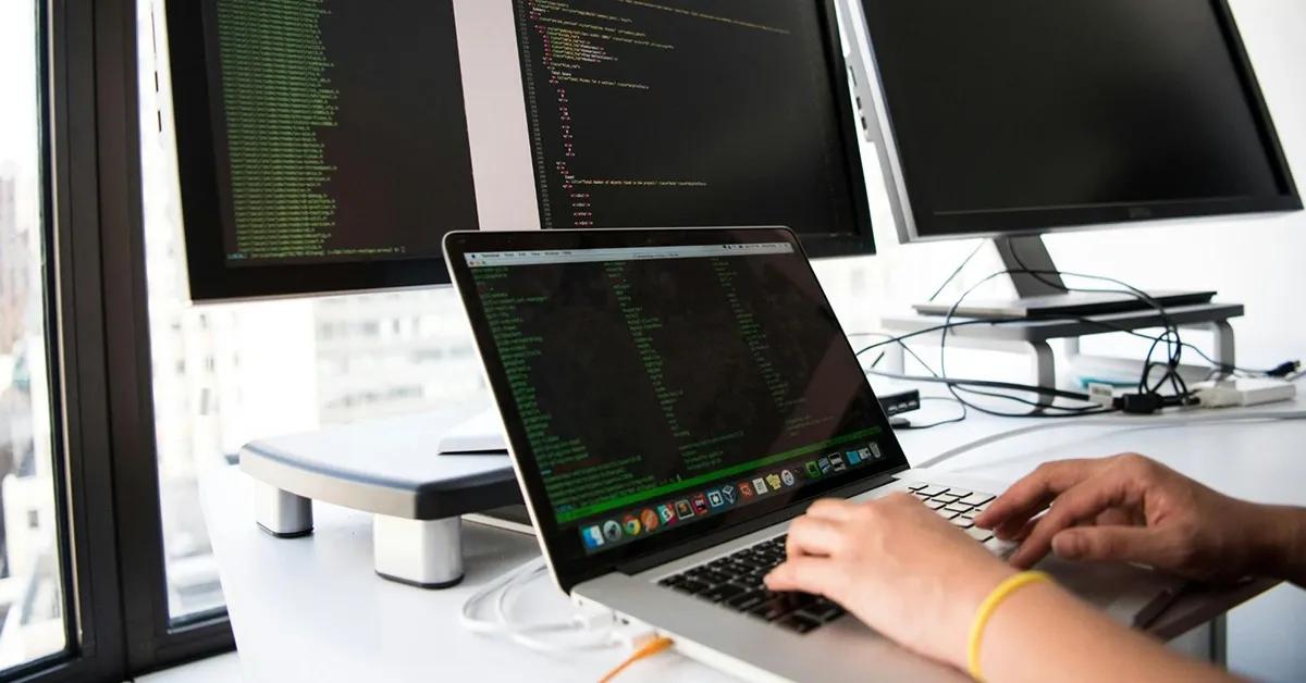 A person typing on their laptop with two desktop computers in backgrounds