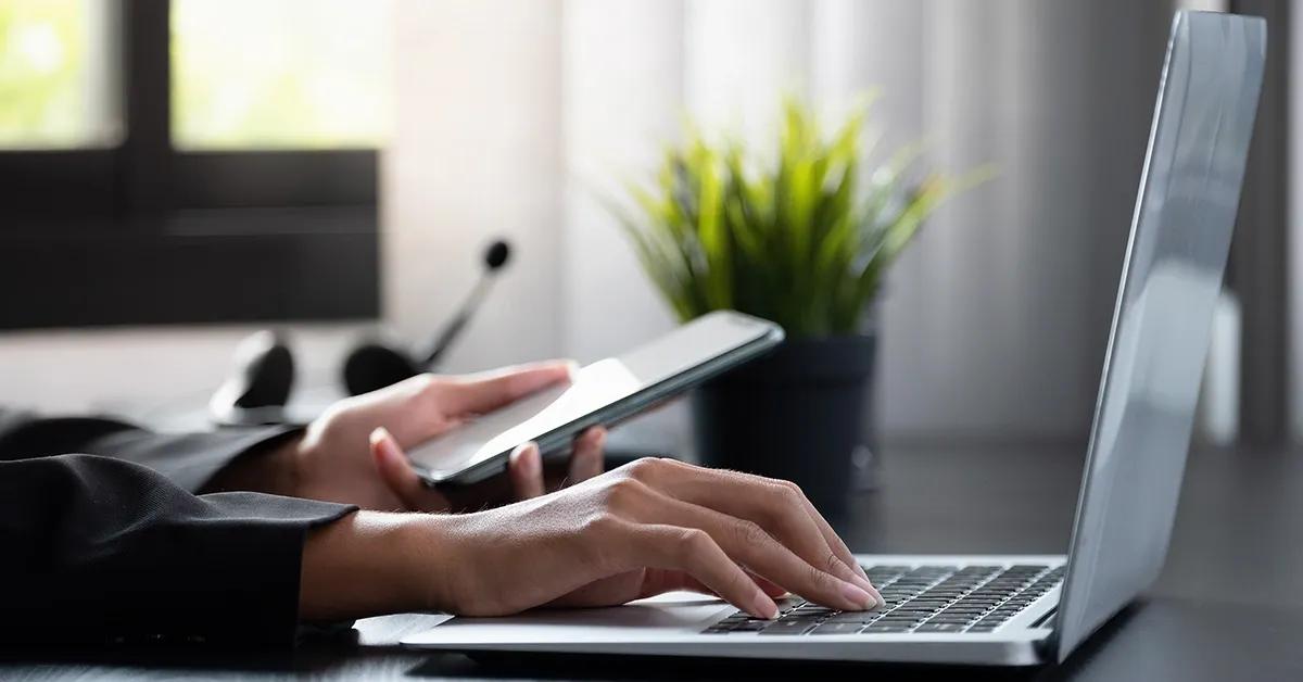 Closeup of person holding a smartphone in one hand and typing on a laptop with the other