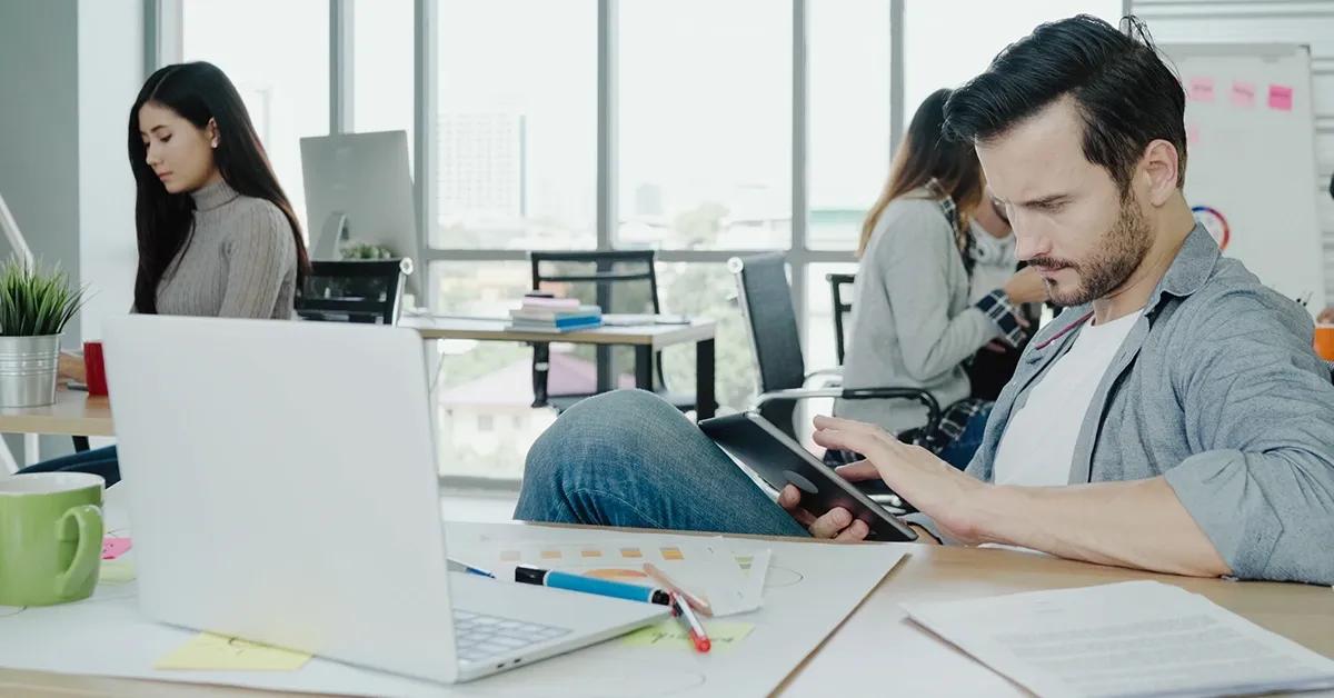 QA engineer tapping on tablet while sitting at a desk in an office and facing a laptop computer