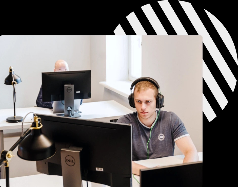 Two people working at desks with large monitors, one wearing headphones, focusing on their screens.