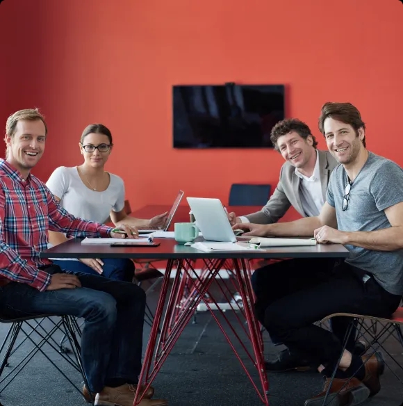 Four team members sitting at a table in a meeting room, all smiling and looking directly at the camera while working on laptops.