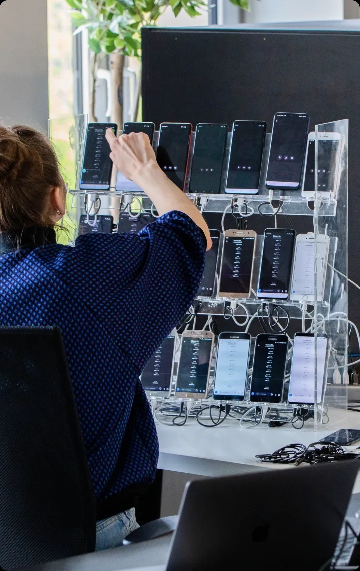 Ingeniero de QA eligiendo un teléfono móvil entre muchos dispositivos de prueba en un soporte.