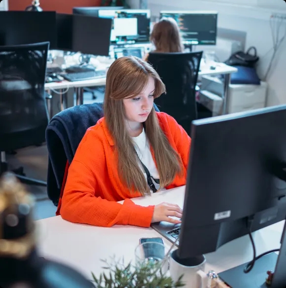 Un ingeniero de QA trabajando en un escritorio de oficina con un portátil en uso y varios monitores visibles en el fondo.