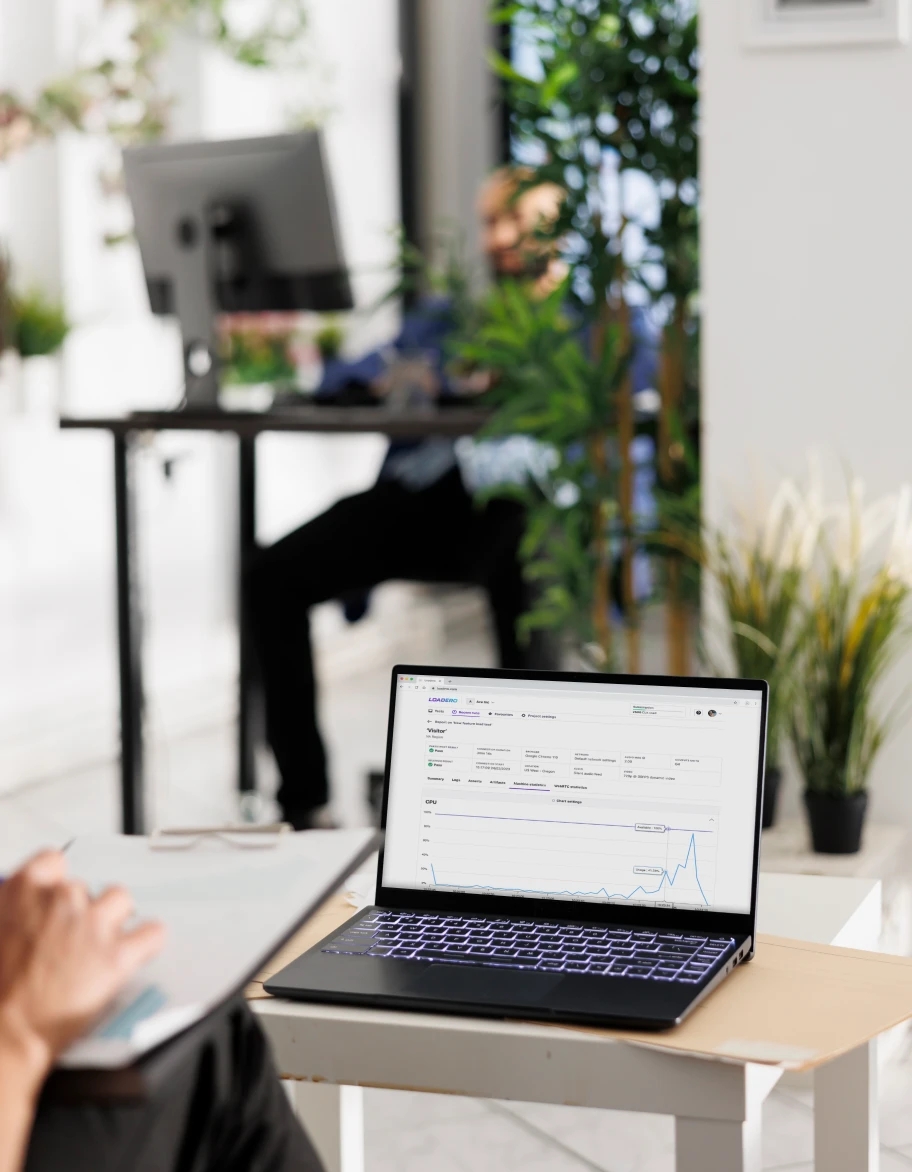 Un espace de bureau ouvert avec des plantes, comprenant un ordinateur portable sur un petit bureau affichant des graphiques de données, avec une personne travaillant en arrière-plan.