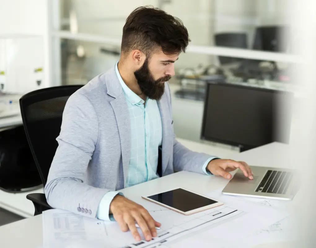 A person in a blazer working at an office desk with a laptop, a tablet and printed documents