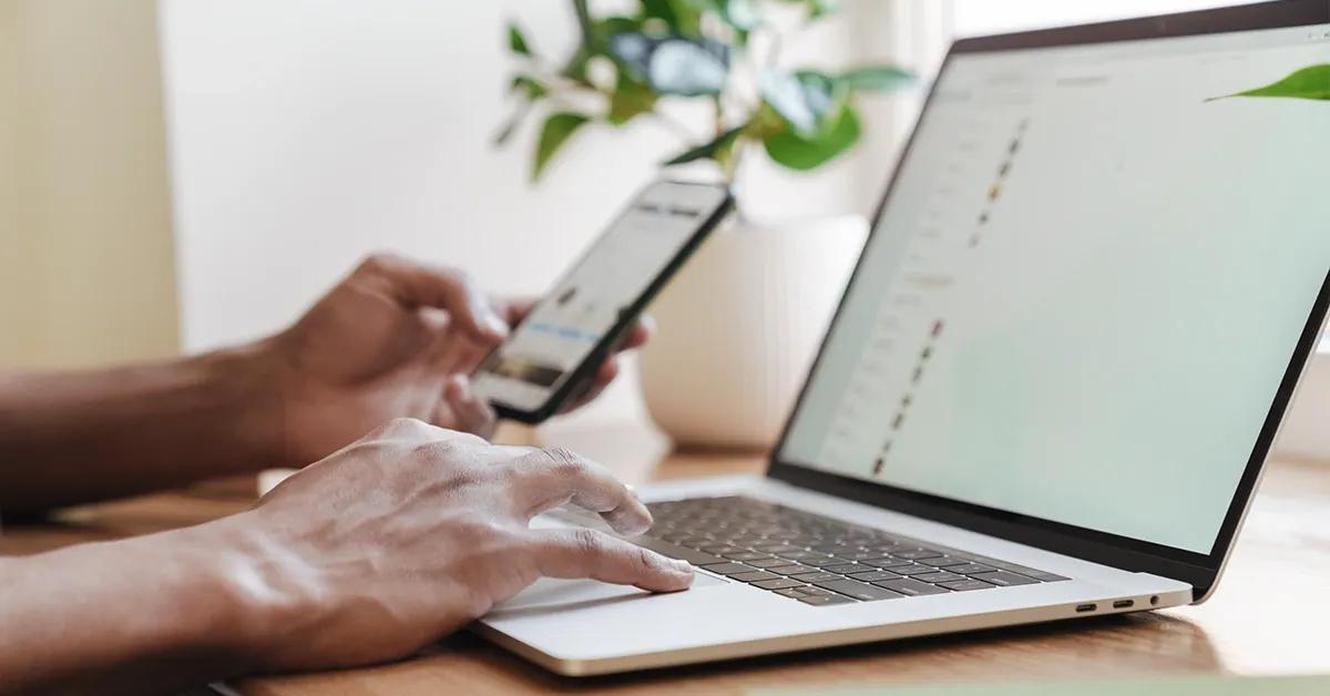 Person typing on laptop and holding a phone