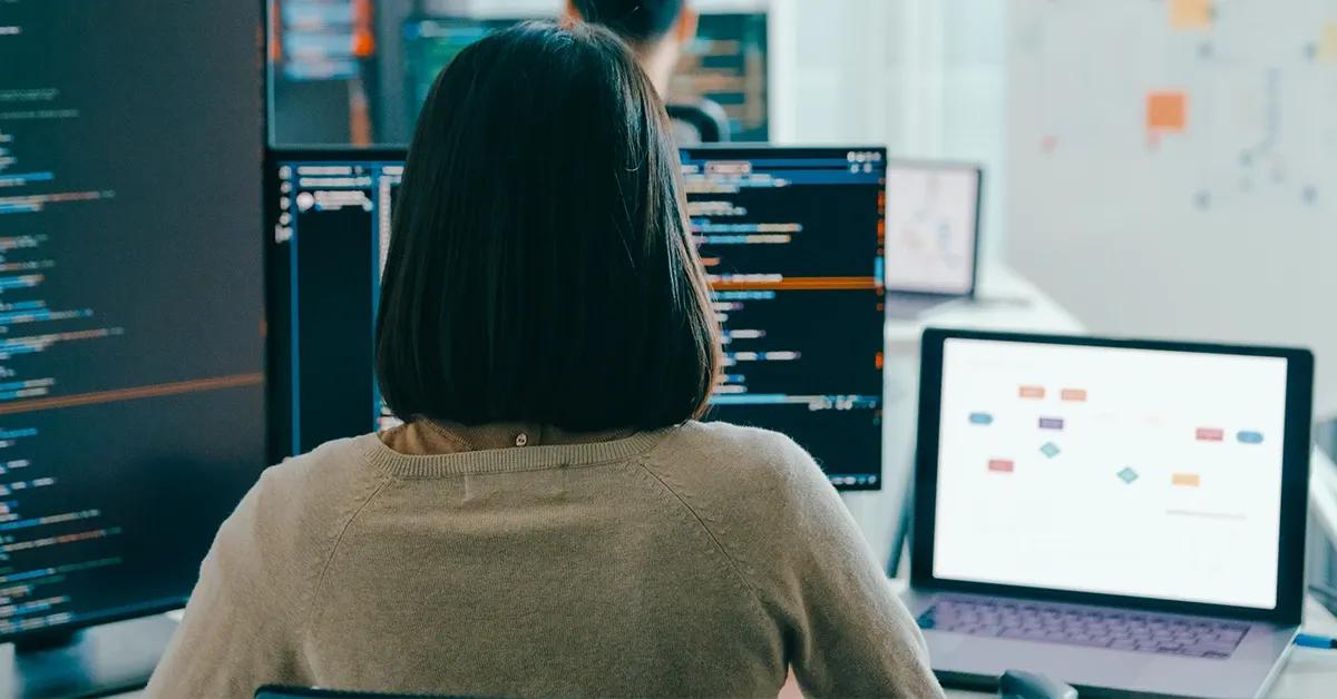 QA engineer sitting at desk with back turned facing multiple screens and devices