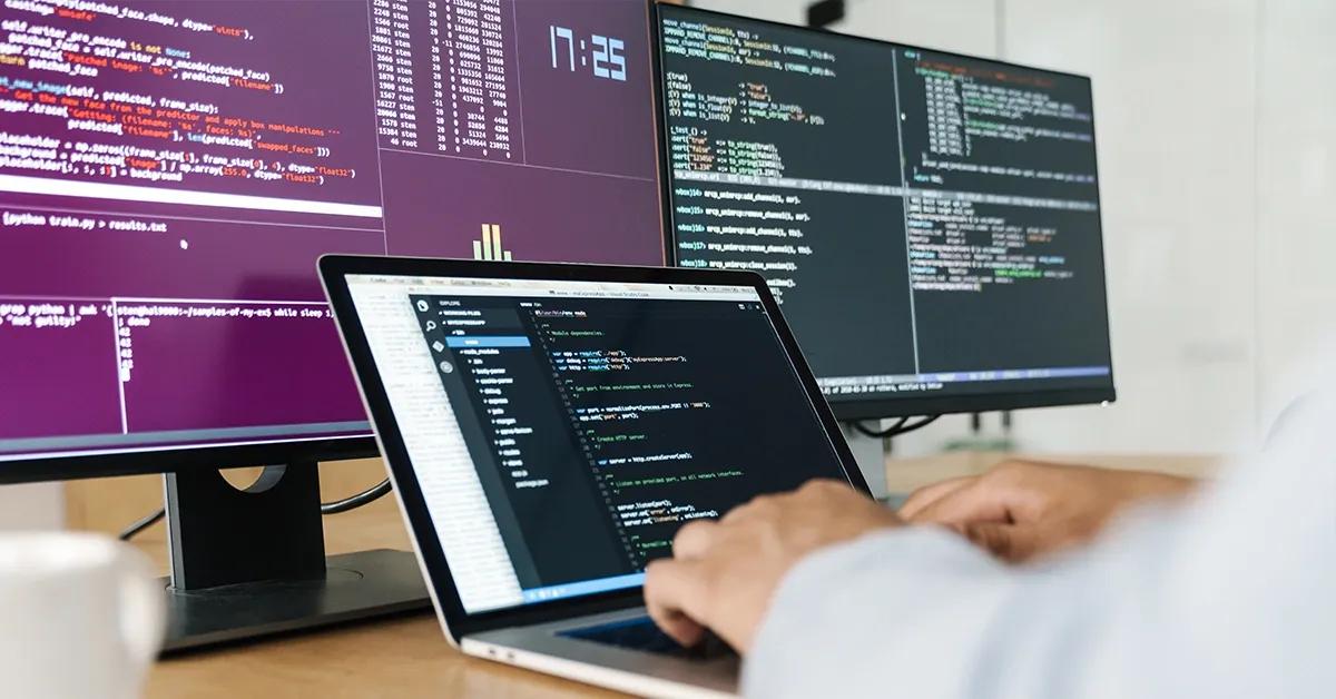 Software engineer writing code on laptop with code visible on two desktop screens in the back