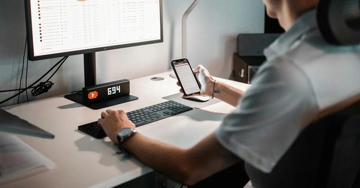 Man sitting at desk holding a smartphone and working on a desktop computer