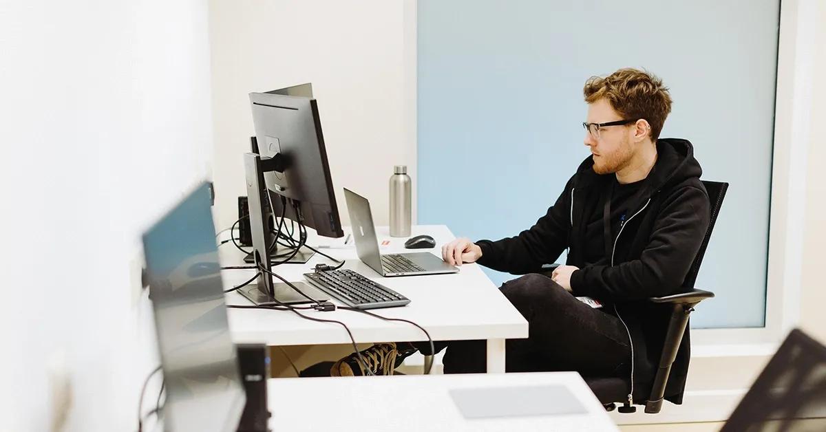 Man sitting at desk working on laptop and facing two desktop computers