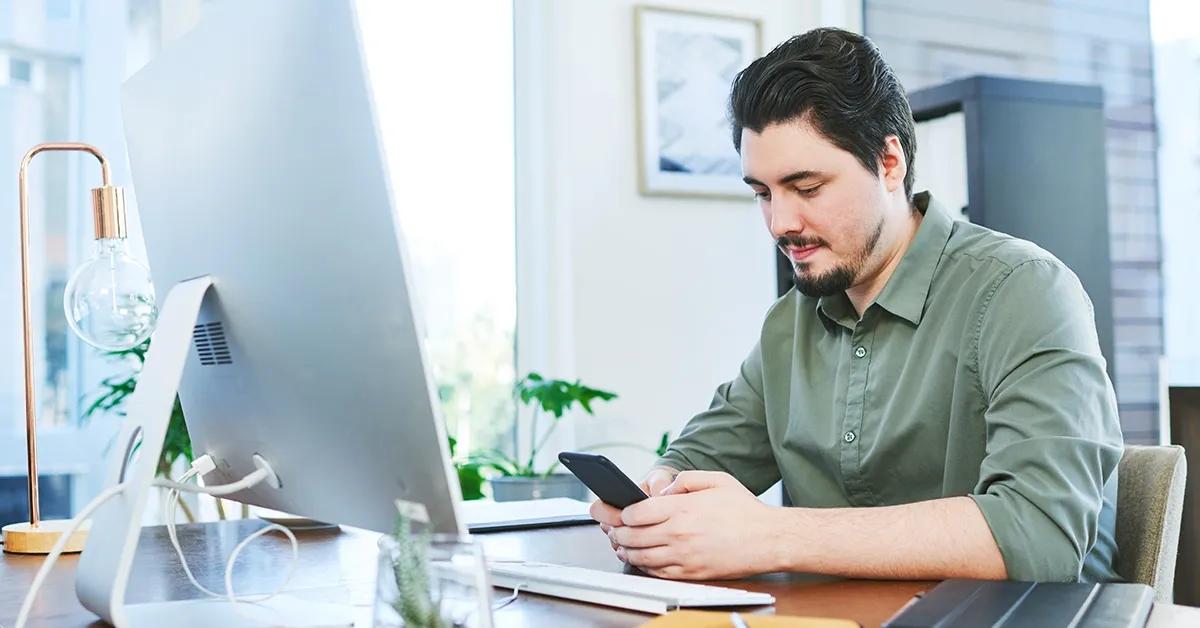 QA engineer sitting at a desk facing a desktop computer while interacting with mobile phone