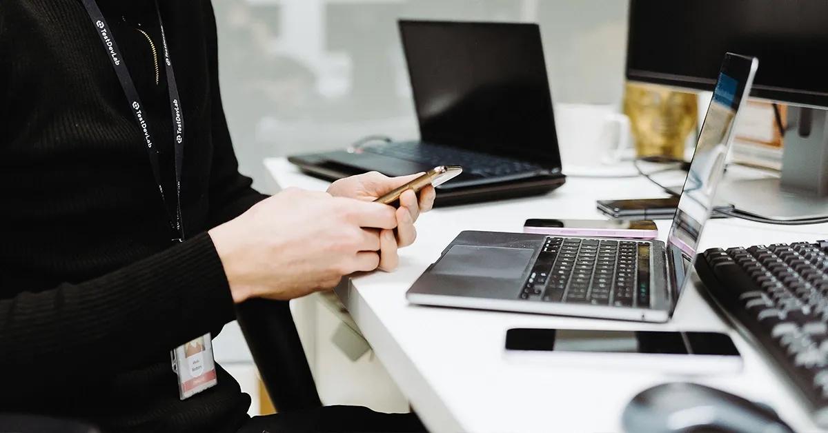 QA engineer sitting at desk with various devices and holding a smartphone