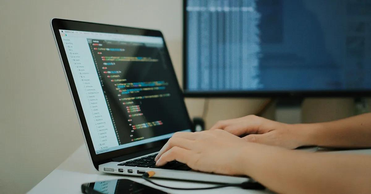 Person writing code on laptop with a desktop computer in background